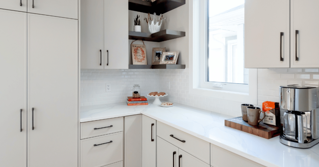 A modern butler’s pantry with white cabinets, white quartz countertops, and a bright window on the right hand side.